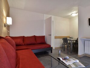 A compact room with a red sofa, a small table with a magazine, a dining area with chairs, and a kitchen area with a refrigerator.