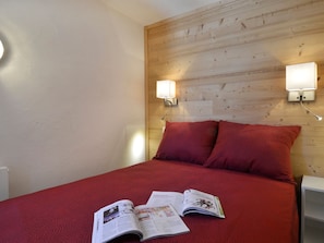 Quarto com cabeceira de madeira, duas luzes de leitura fixadas na parede, colcha vermelha e uma revista sobre a cama.