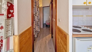Un pasillo estrecho con suelo de madera que conduce a un dormitorio con cama y mesita de noche. A la derecha se ve una zona de cocina con estufa y armarios.