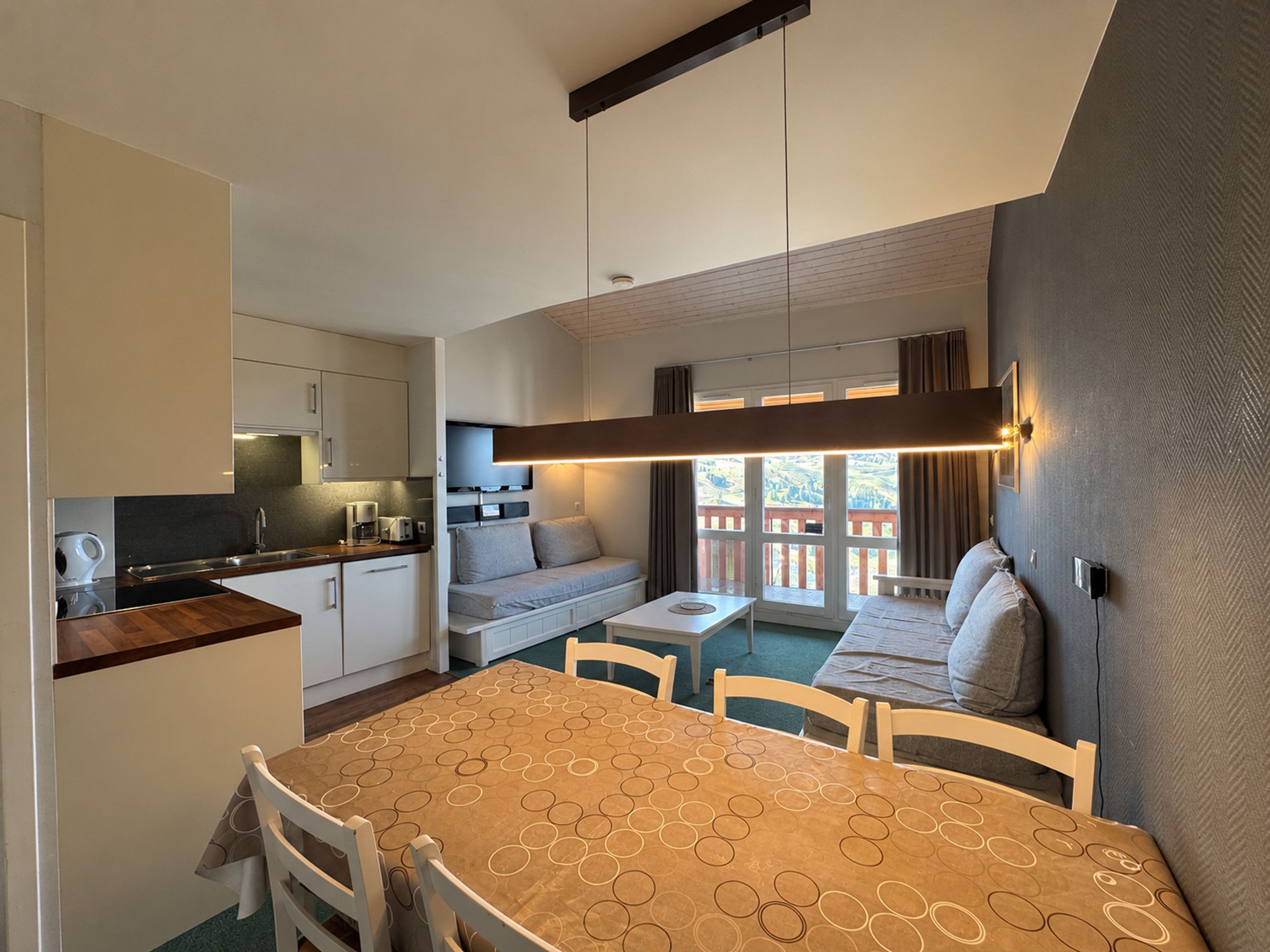 A compact kitchen with modern appliances, a dining area with a patterned tablecloth, and a balcony with a view.