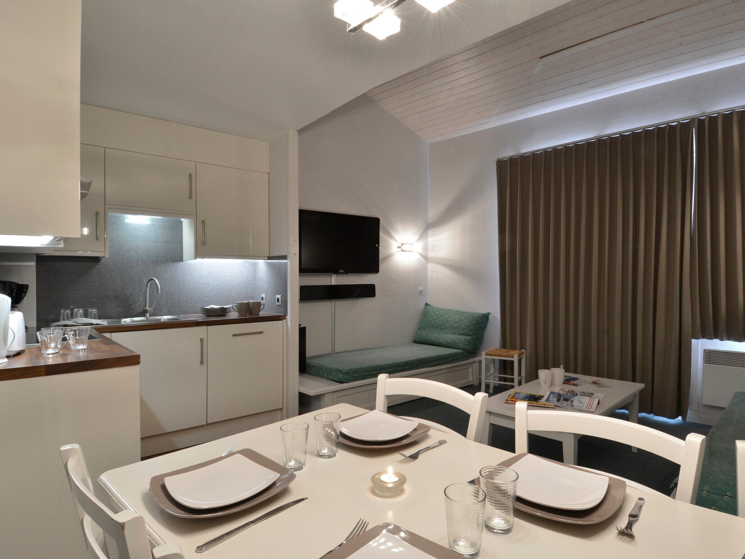 Une chambre d’hôtel compacte comprenant un coin cuisine, un espace repas, un téléviseur fixé au mur et une grande fenêtre avec des rideaux.