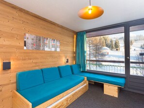 A wooden interior with a blue sofa, a wall-mounted light, and a window with a view of a snowy landscape. - Renovated Studio 5 ppl · Ski-in/Ski-out · Balcony · Pets Allowed (Les Arcs 1800)