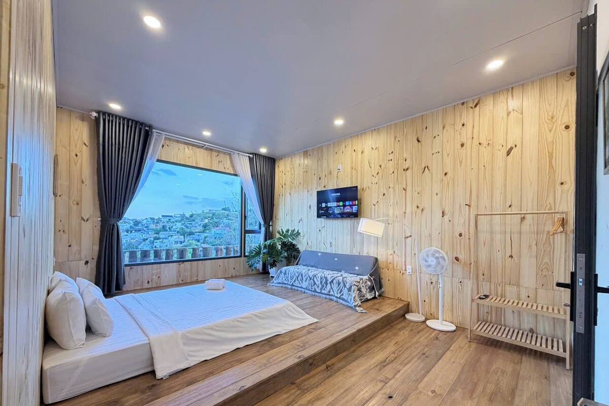 Une chambre à coucher avec un lit, une télévision, une fenêtre avec des rideaux, une plante et un ventilateur.