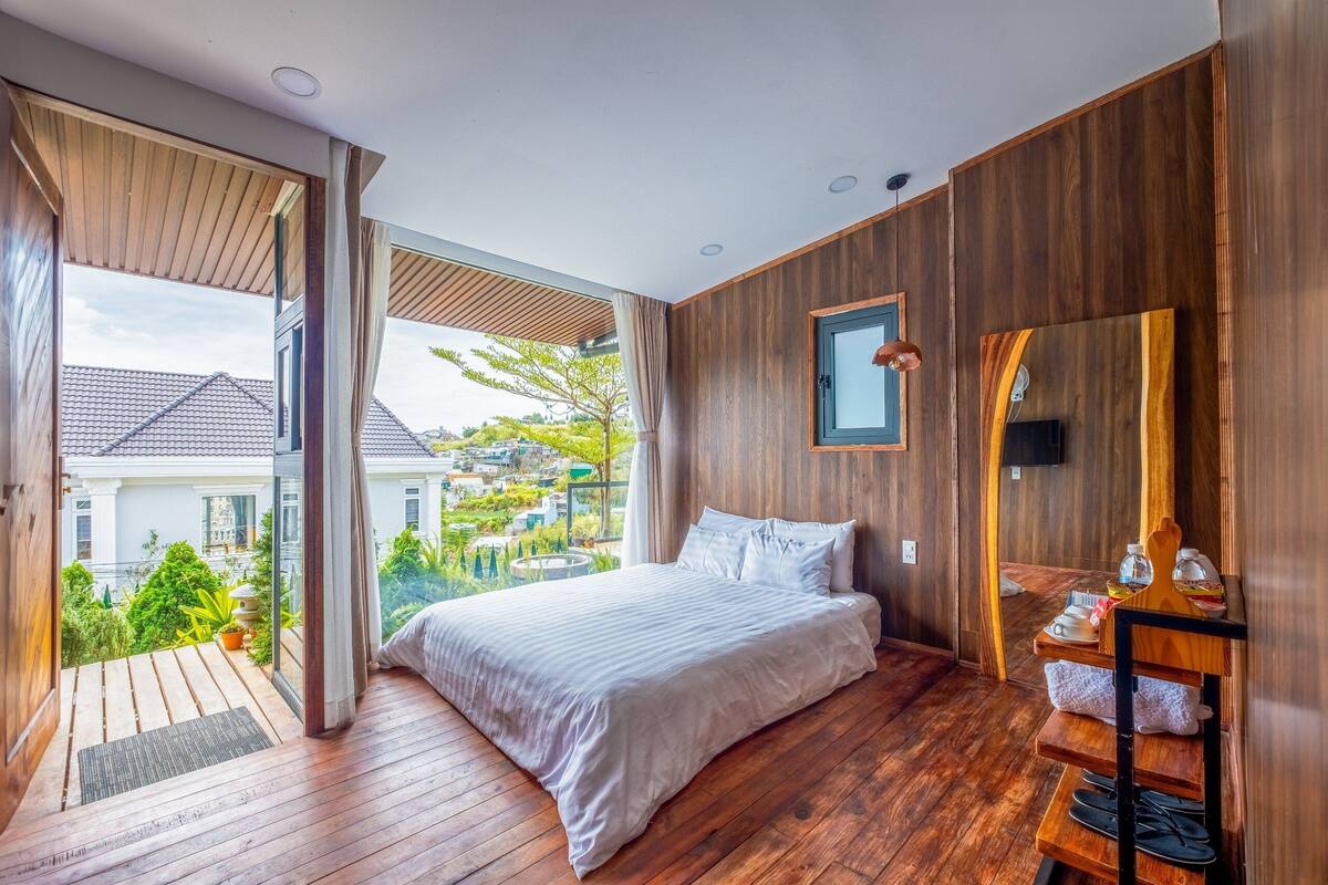 Une chambre avec un grand lit, un plancher en bois, un plafond en bois, une vue sur la verdure et une petite étagère en bois sur laquelle sont posés quelques objets.
