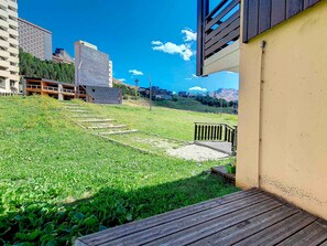 Uma área externa gramada com um deck de madeira, um caminho de pedra e vista para edifícios ao fundo.