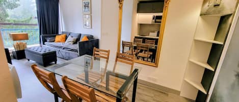 Un salon moderne avec une table à manger en verre, des chaises en bois, un canapé avec des coussins, un grand miroir et une vue sur le balcon.