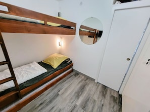Uma cama beliche com estrutura de madeira, um colchão individual e um travesseiro amarelo. Há um espelho redondo na parede e um guarda-roupa branco com porta.