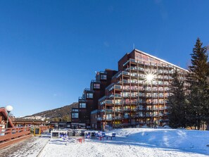 Um hotel com vários andares e varandas, com uma pista de esqui em primeiro plano e montanhas ao fundo.