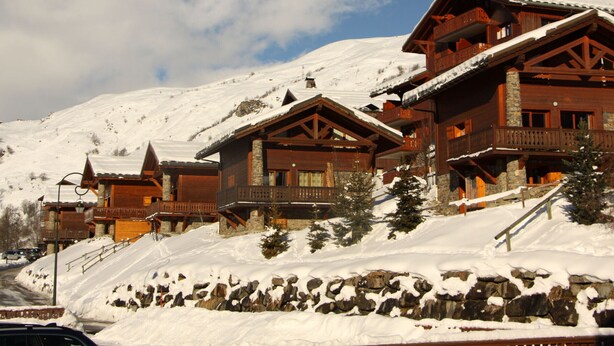 A wooden lodge with multiple balconies and a stone foundation, surrounded by snow-covered terrain and a clear sky. - Spacious apartment near slopes with balcony for 8 people (Les Menuires)