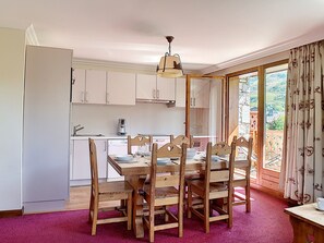 Uma zona de refeições com uma mesa e cadeiras de madeira, uma cozinha ao fundo e vista para o exterior através de uma porta de vidro.