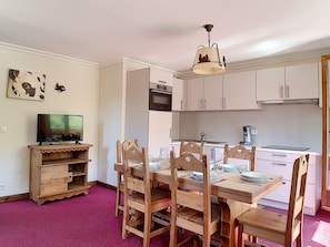 Uma sala de jantar com uma mesa de madeira posta para quatro pessoas, uma televisão sobre um suporte de madeira e uma cozinha ao fundo.