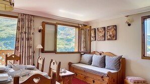 A cozy room with a wooden dining table set for four, a bench with cushions, and a large window with a view of trees. - Renovated 3-room apartment near slopes, balcony, sauna, steam room, WIFI (Les Menuires)