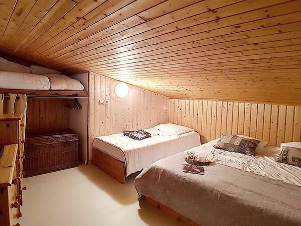Ein gemütliches Schlafzimmer im Dachgeschoss mit Holzwänden und schrägen Decken. Es gibt zwei Betten, ein Etagenbett und einen kleinen Schreibtisch mit Stuhl.