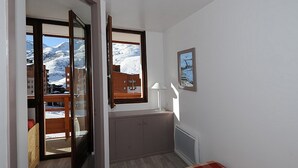 A room with a bed, a window with a view of snow-covered mountains, a small table with a lamp, and a framed picture on the wall. - Renovated 2-room apartment, ski-in/ski-out, southwest balcony (Les Menuires)