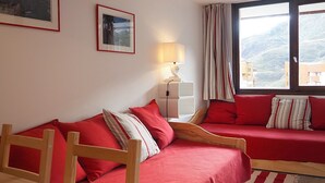 A room with two red beds, a wooden chair, a lamp, and a window with a mountain view. - Renovated 2-room apartment, ski-in/ski-out, southwest balcony (Les Menuires)