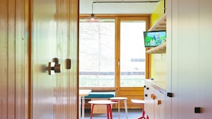 A room with a wooden door, a window with a view of a balcony, a table with red chairs, and a television on a shelf.