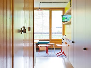 Una habitación con puerta de madera, ventanal con vistas, mesa con sillas y televisor sobre una estantería.