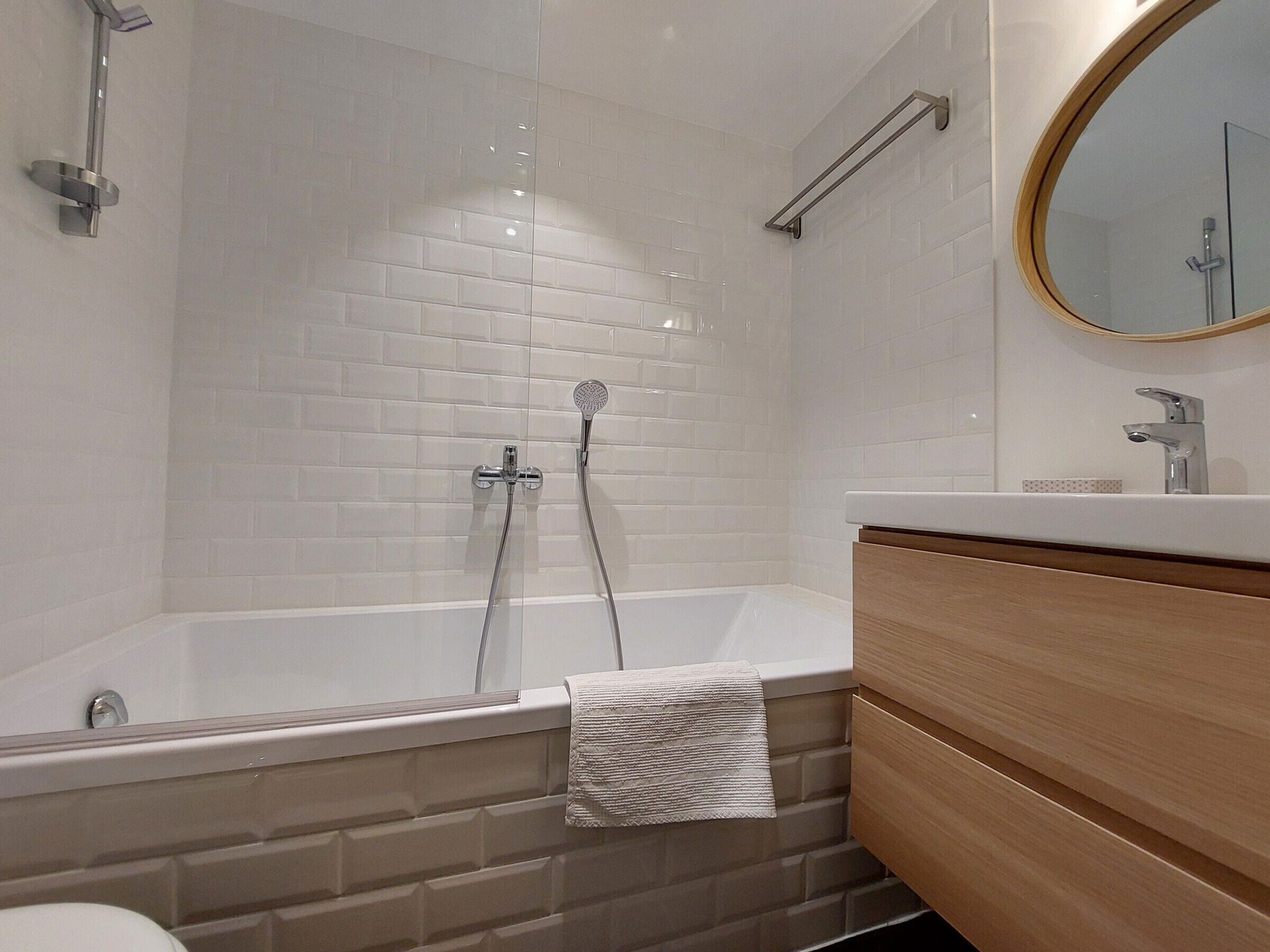 Un bagno moderno con doccia rivestita di piastrelle bianche, un mobile lavabo in legno e uno specchio rotondo.