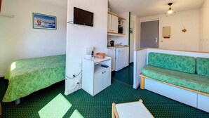 Ein kleines Zimmer mit zwei Einzelbetten, einer Küchenzeile, einem grünen Sofa und einem Fernseher.