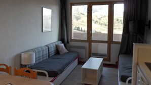 Una habitación pequeña y escasamente amoblada con un sofá, una mesita y una ventana con vistas a un paisaje montañoso.