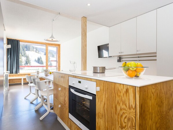 A modern kitchen with wooden cabinets, a built-in oven, a large window with a view of a snowy landscape, and a bowl of fruit on the counter.