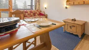 Een gezellige kamer met een houten eettafel, een bank met een sprei, een klein houten kastje en een raam met uitzicht op bomen.