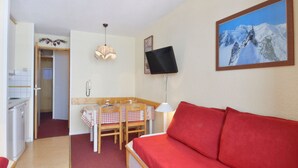 Een gezellige kamer met een rode bank, een eettafel met stoelen, een keukenhoek en een schilderij van een besneeuwde berg.