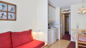 Een knus ingerichte kamer met een rode bank, een kleine kitchenette, een houten tafel met geruit tafelkleed en een wandlamp.