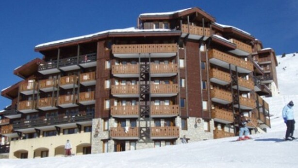 Een hotel met meerdere verdiepingen, balkons, skipistes en een helderblauwe lucht.