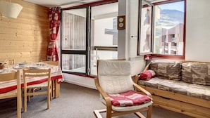 A cozy room with a dining table set for four, a cushioned sofa, a wooden chair, and a large window offering a view of buildings and hills. - Spacious apartment 6 pers. Near slopes with parking (Les Menuires)