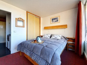 A bedroom with a bed, a bedside table, a nightstand, a window with red curtains, and a picture on the wall.