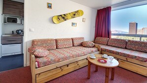 A compact hotel room with a sofa set, a small table, a microwave, and a view of a cityscape through a window. - Cozy apartment for 4 with balcony at ski slopes (Les Menuires)