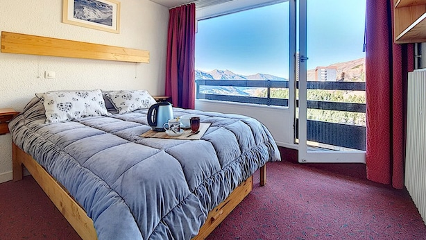 A bedroom with a bed, a window with a view of mountains, a wooden headboard, and a red carpet. - Cozy apartment for 4 with balcony at ski slopes (Les Menuires)