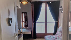 A small, cozy room with a bed, a desk, and a chair. There is a window with curtains, a door, and a wall-mounted light fixture.