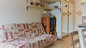 A cozy living room with a patterned sofa, wooden shelves with decorative items, a small cabinet with a lamp, and a wardrobe with a mirror.