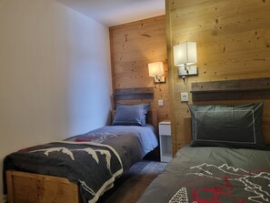 Une petite pièce chaleureuse aux murs en bois, avec un lit recouvert d’une couverture et agrémenté d’oreillers. On y trouve une table de chevet avec une lampe, ainsi qu’une petite table.