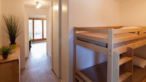 Una cama individual en literas en una habitación con piso de madera, una planta en maceta y una puerta que da a otra habitación.