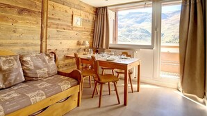 A cozy room with a wooden wall, a wooden sofa with patterned cushions, a dining table set for four, and a large window with a mountain view. - Comfortable duplex 5 pers · Ski-in/Ski-out · Balcony (Les Menuires)