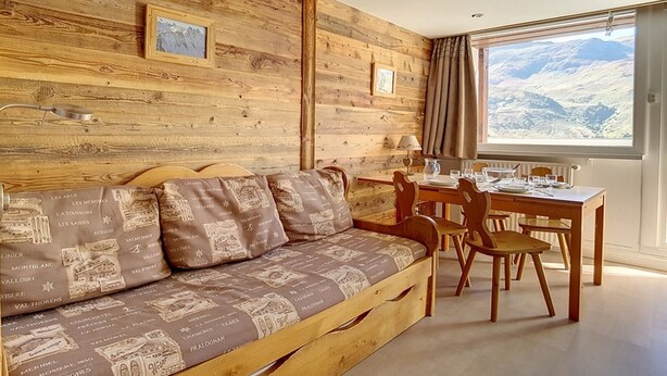 A cozy room with a wooden wall, a sofa with patterned cushions, a dining table with chairs, and a window with a mountain view. - Comfortable duplex 5 pers · Ski-in/Ski-out · Balcony (Les Menuires)
