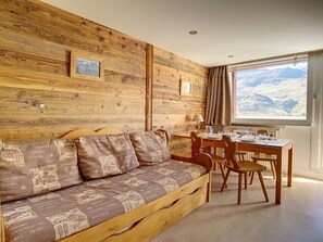 Ein gemütliches Zimmer mit einer Holzwand, einem Sofa mit gemusterten Kissen, einem Esstisch mit Stühlen und einem Fenster mit Bergblick.