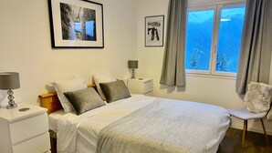 Un dormitorio con una cama, dos mesitas de noche, una silla, un ventanal con cortinas y dos cuadros enmarcados en la pared.