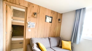 A room with a grey sofa, wooden panel walls, a window with curtains, and a bunk bed with a wooden door. - Studio cabin at the slopes, pets allowed (Tignes)