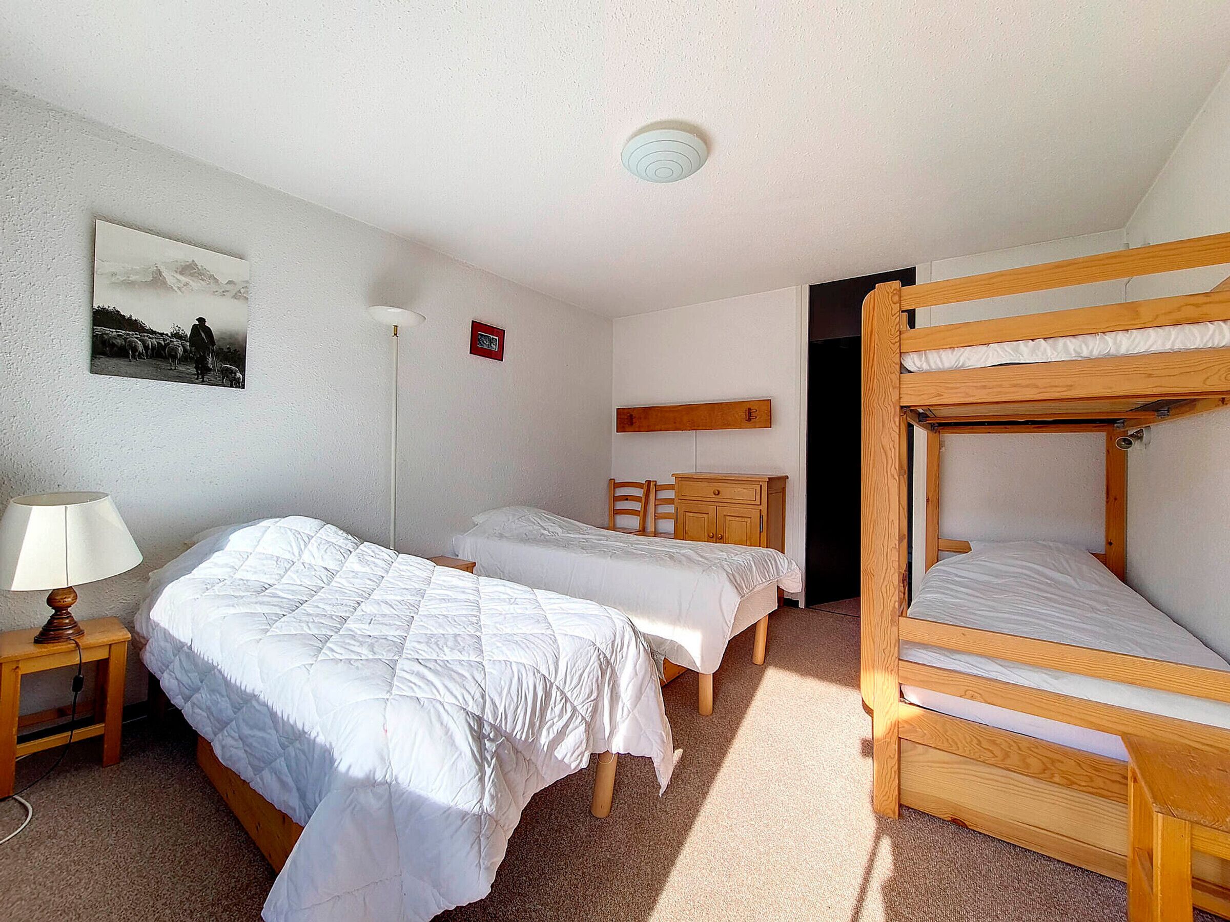 Una habitación con dos camas individuales, una litera, un escritorio, una silla, una lámpara y un cuadro en la pared.
