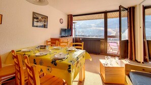 A dining area with a table set for four, a television on a wooden cabinet, and a balcony with a view of the landscape. - Bright 2-Room Apartment - Ski-in/Ski-out - Balcony - 6 ppl - Wifi (Les Menuires)