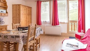 A cozy room with a dining table set for four, wooden cabinets, a red sofa, and large windows with curtains. - Functional studio, ski-in/ski-out, balcony (Les Menuires)