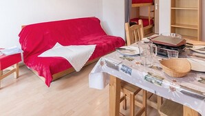 A cozy room with a red sofa, a dining table with a floral tablecloth, and a wooden bookshelf. - Functional studio, ski-in/ski-out, balcony (Les Menuires)