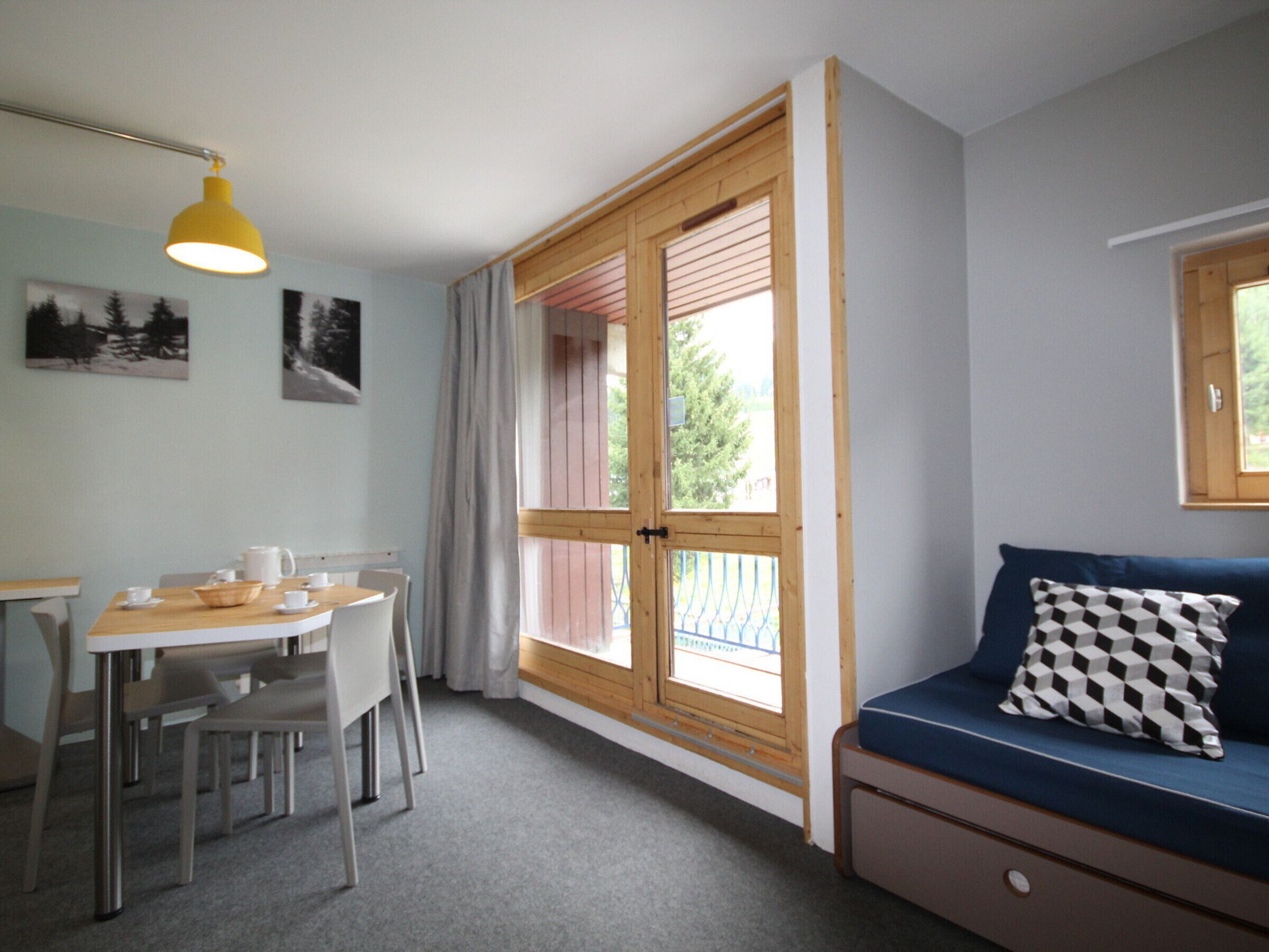 Een kamer met een eettafel, stoelen, een bank met een geruit kussen, een raam met uitzicht op bomen, en een gele hanglamp.