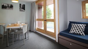 A room with a dining table, chairs, a sofa with a checkered pillow, a window with a view of trees, and a yellow pendant light.