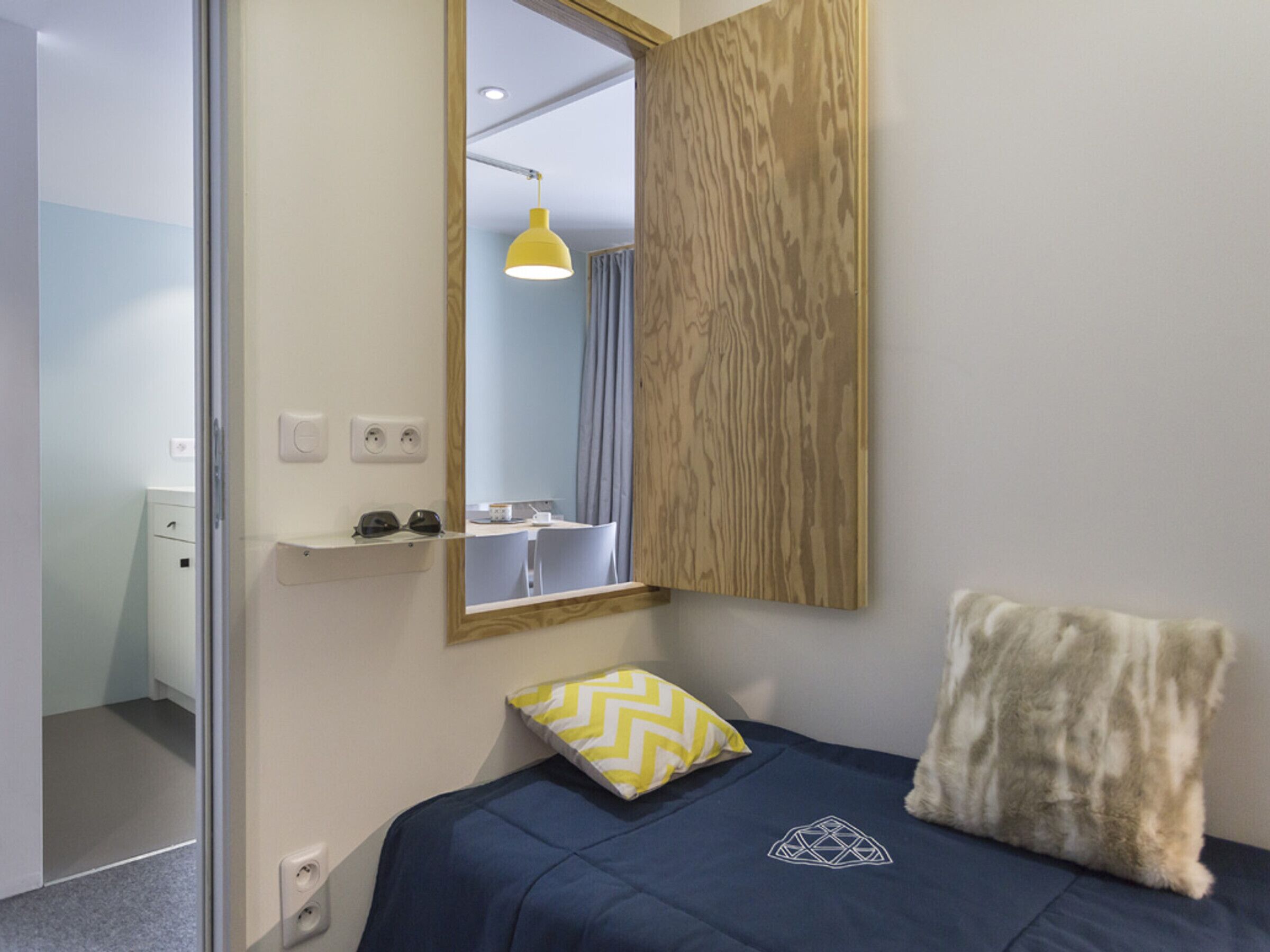 Ein Schlafzimmer mit Bett, Spiegel, einer Holzwand, einer gelben Pendelleuchte und einem kleinen Schreibtisch mit Stuhl.