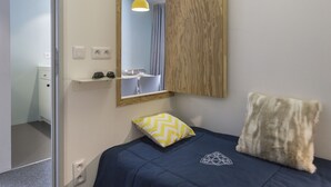 A bedroom with a bed, a mirror, a wooden panel, a yellow pendant light, and a small desk with a chair.